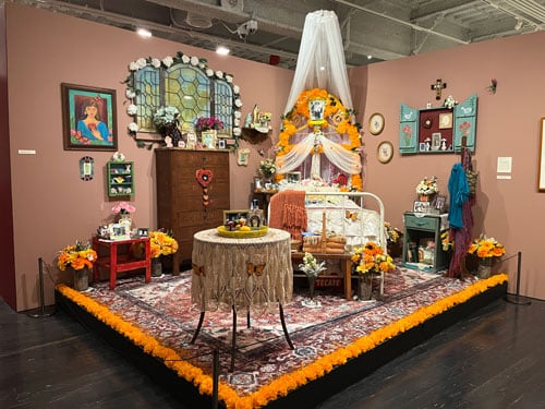 An installation view of a work by Ofelia Esparza shows a woman's bedroom that has been recreated in a museum gallery and trimmed with bright orange flowers.