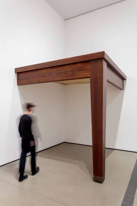 A man walks under a monumental sculpture of a table tucked into a corner.