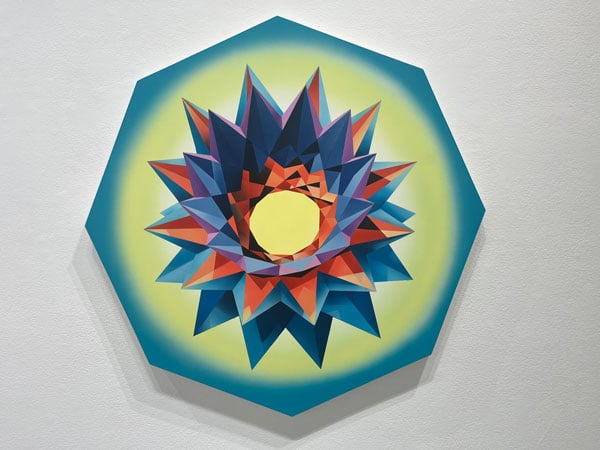 A painting by Mary Anna Pomonis on a hexagonal canvas features a colorful geometric pattern resembling a flower in shades of blue, red and a very light yellow.