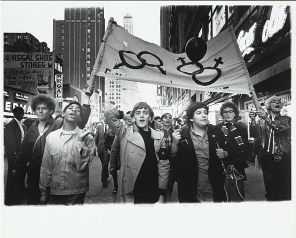 A vintage black and white image shows protestors holding a sign with gay rights symbols.