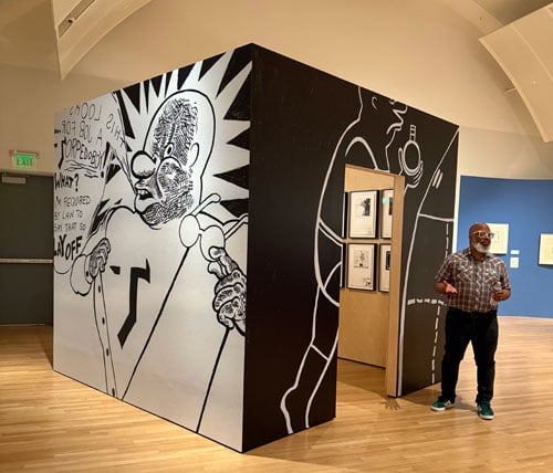 Artist Trenton Doyle Hancock, a Black man with a white beard, stands before a cube illustrated with a cartoonish Black figure called Torpedoboy (his alter ego).