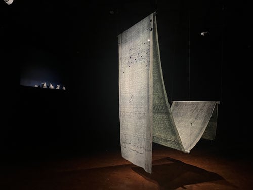 An installation by Swapnaa Tamhane shows a long textile with a faint blue geometric pattner hanging in a dramatically illuminated gallery.