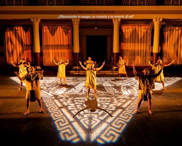 A color photograph shows seven figures in masks singing, with their arms extended, before a classical facade. A Mesoamerican pattern is projected onto the floor below them.