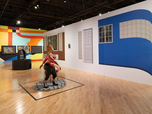 An installation view of "Dear Mazie" shows a wall bearing arquitectonic vinyls that reveal elements of a house, including a blue wall with glass blocks and a doorway. At left, a sculpture by Tschabalala Self depicts a curvy Black woman in a red top and Black pants.