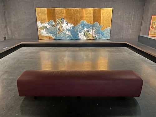 A long, upholstered bench stands before a Japanese folding screen showing stylized waves against a gold background.