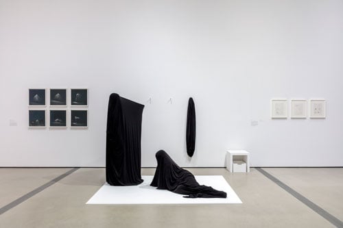 A gallery features two people wearing black bags, standing on shallow white platform. One person stands, the other rests on their side.