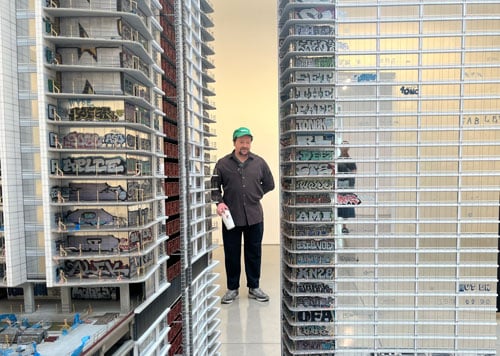 Artist Sayre Gomez, wearing a dark button down shirt and green baseball cap, is seen standing between portions of a sculpture that show corporate towers covered in graffiti.