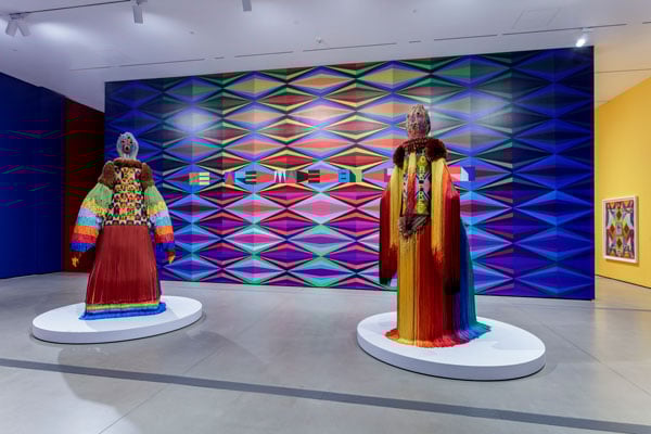 A gallery covered in brightly patterned geometric wallpaper features a pair of totemic figures in colorful costume in the foreground.