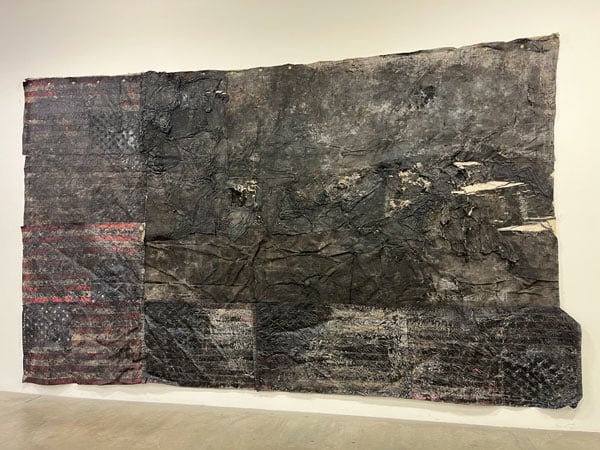 A work of art by Molly Tierney hangs on a wall, showing U.S. flags collaged together, and covered by an inky black substance — looking as if the flags had just burned.