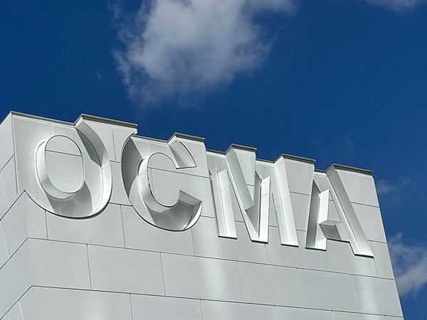 A close-up shows the logo of the Orange County Museum of Art — OCMA — against the roofline of its all-white building.