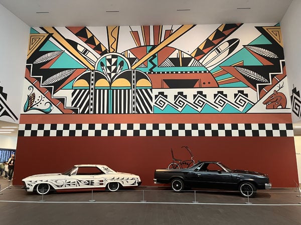 Two lowriders bearing Tewa pottery designs are parked before a brilliant Pueblo mural in a museum gallery.