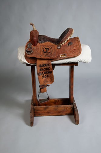 A color photograph shows a natural leather, hand tooled saddle resting on a wooden saddle rack.