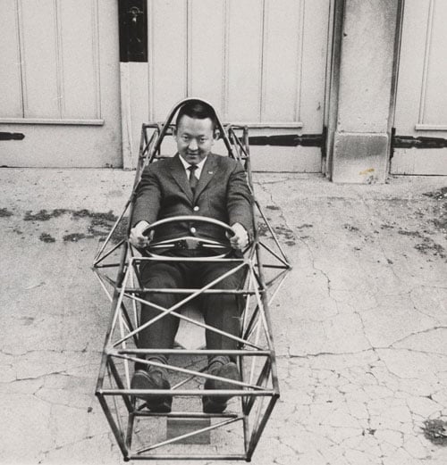 A Japanese American man in a suit is seen posing in the bare steel frame of a hot rod.