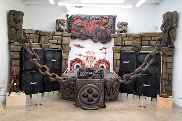 An installation in a gallery bears a large sign that reads GWAR hanging over two gargoyles bearing large chain and a drum set that looks as if it were lined with meat.