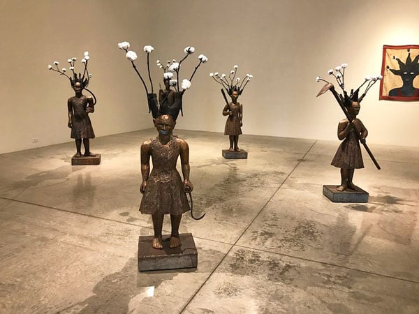 An installation view of a gallery space shows a series of Black female figures bearing agricultural tools who bear spiky hair that morphs in pieces of cotton plant.