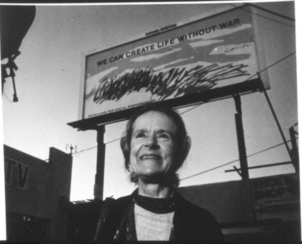 A black and white photo shows Corita Kent standing before a billboard that reads "We Can Create Life Without War"
