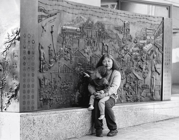 A Japanese American woman sits with a child on her lap in front of a bas relief monument.