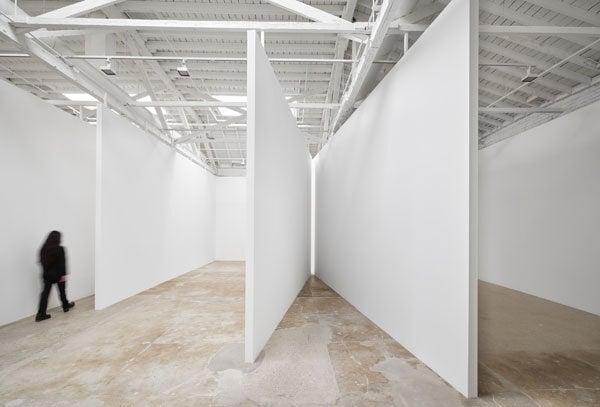An installation view of a gallery shows a blurry figure walking into a white walled architectonic installation