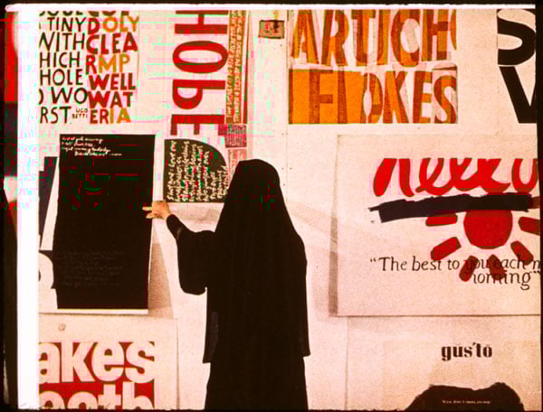A nun is seen adjusting prints with bright color texts against a wall