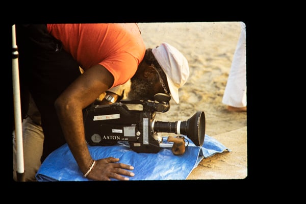 A Black man leans over a film camera during a shoot on a beach