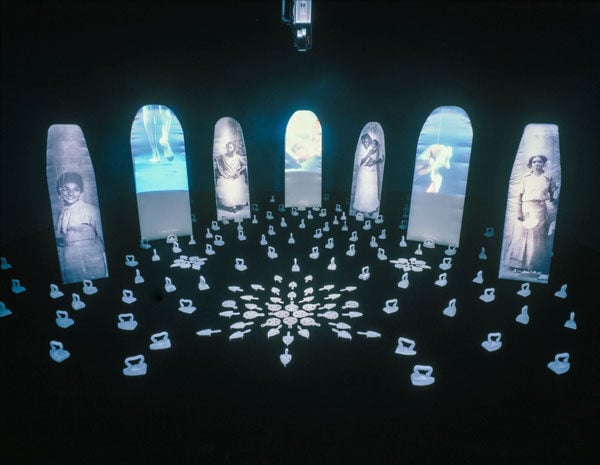 In a dim room, a row of screen shaped like ironing boards are illuminated with images of women. On the floors sits an arrangement of vintage irons.