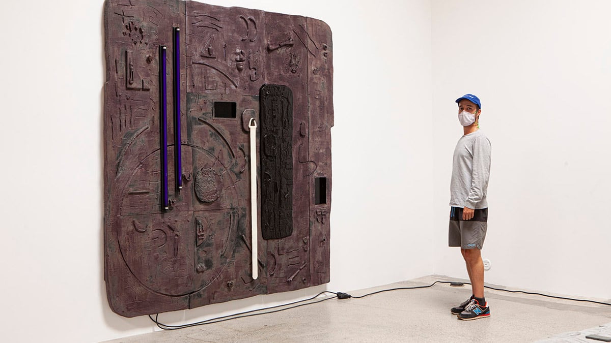 Artist Don Edler with his artwork, “Death Tablet,” 2018. Plywood, OSB, hydrocal, styrofoam, acrylic, platinum silicone, fake bones, barbie doll, black light, fluorescent tube, pine, clear packing plastic, gorilla tape, air conditioner. 120 in x 120 in x 3 in. Image courtesy of the artist. 