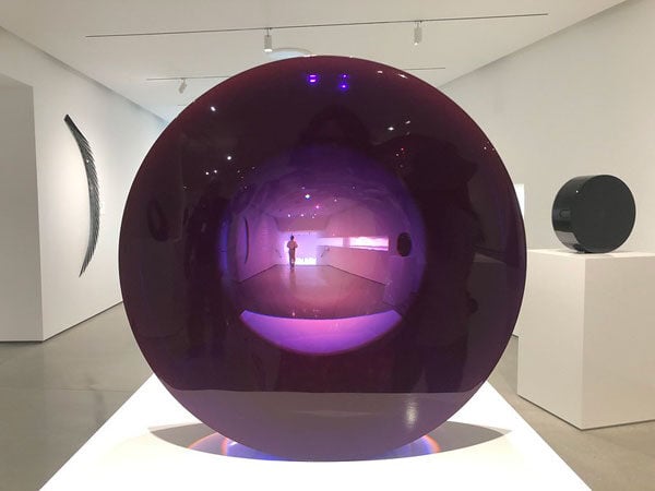 A large purple lens sculpture stands on a white plinth in a gallery.