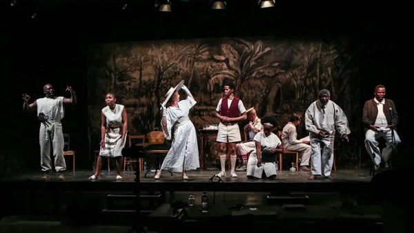 A White woman in an all white ensemble inspects a Black boy in a scene from Houseboy