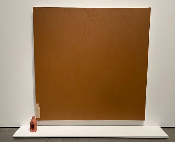 A milk bottle painted the color of terracotta sits before a canvas of the same color