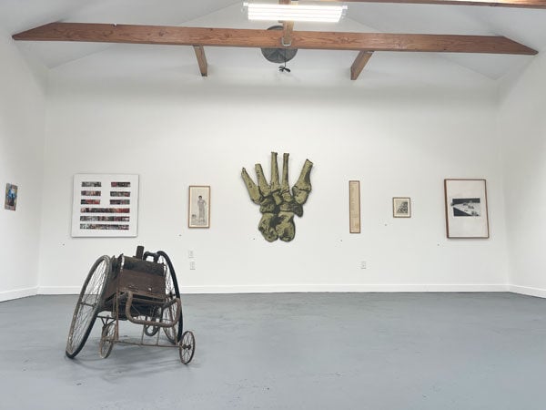 A view of a gallery shows a mechanical sculpture that resembles a wheelchair and paintings hanging on a wall