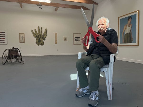 An older man sits in a gallery holding a giant pair of red scissors.