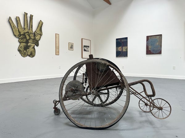 An installation view of a gallery shows a sculpture made of metal that resembles a wheelchair and paintings on a wall.
