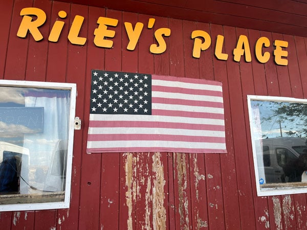 The wooden facade of a bar bears a U.S. flag and a large sign that reads "Riley's Place"