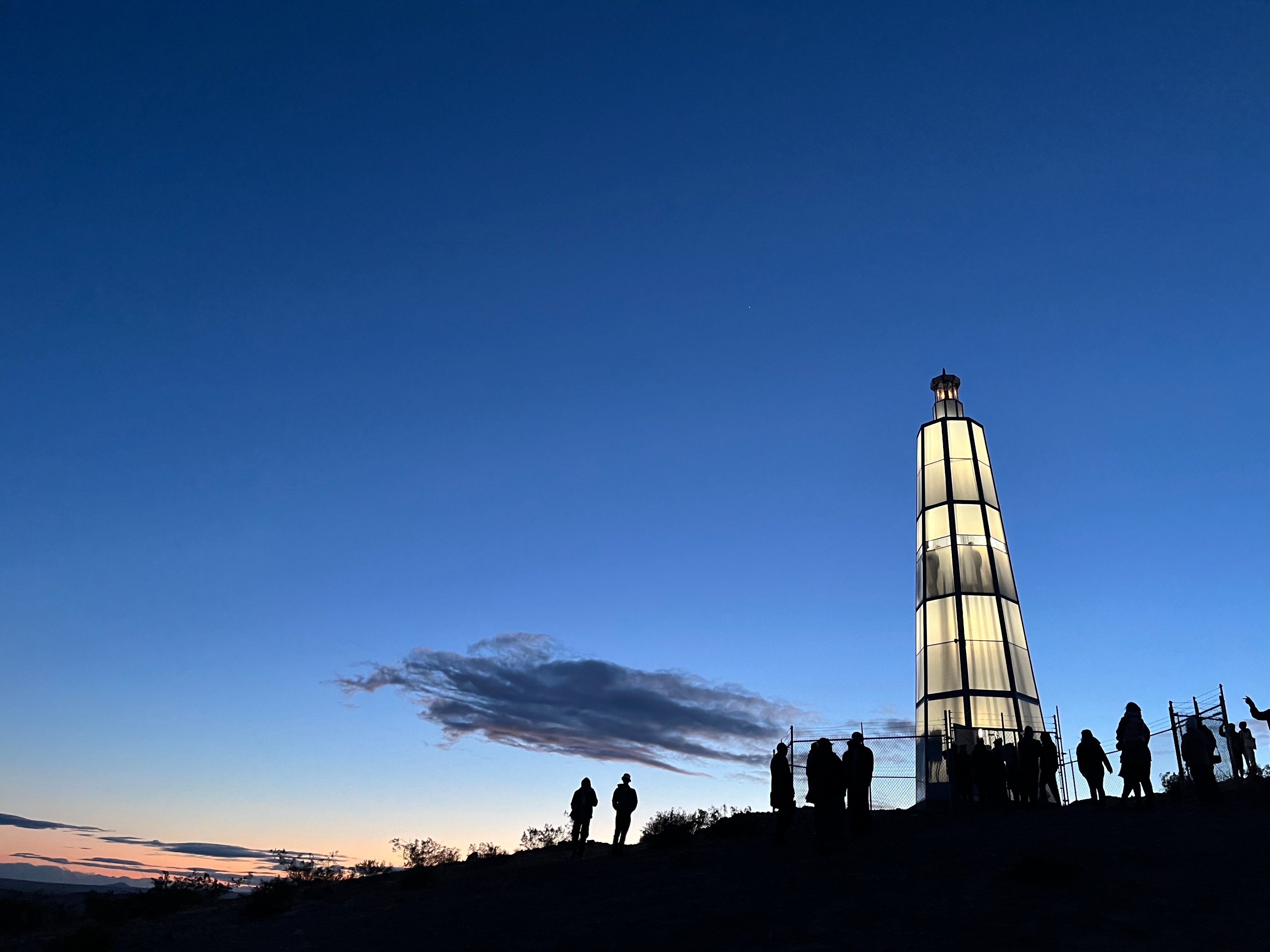 Silhouettes of people can be seen standing before a lighthouse standin gon a small rise at dusk.