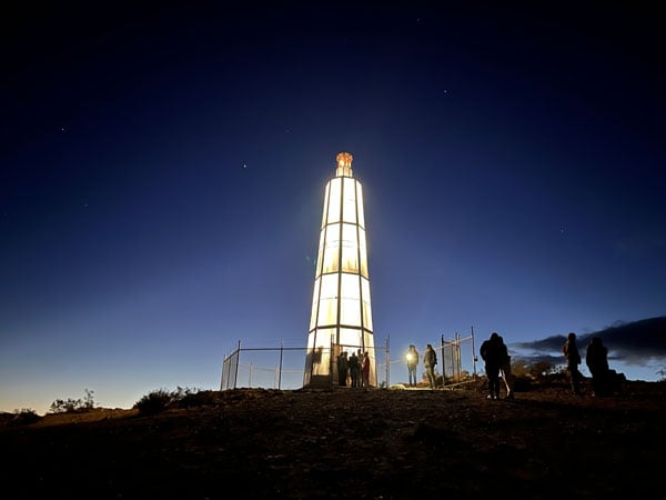 A lighthouse structure, illuminated from within, stands on a windswept hill in the desert