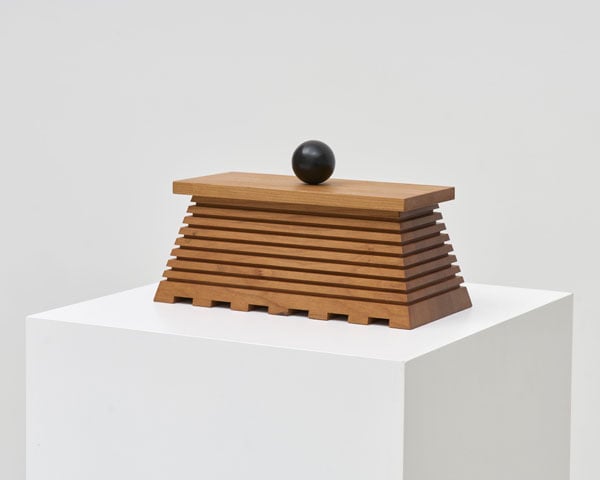 A trapezoidal wooden box rests on a white plinth in a gallery.