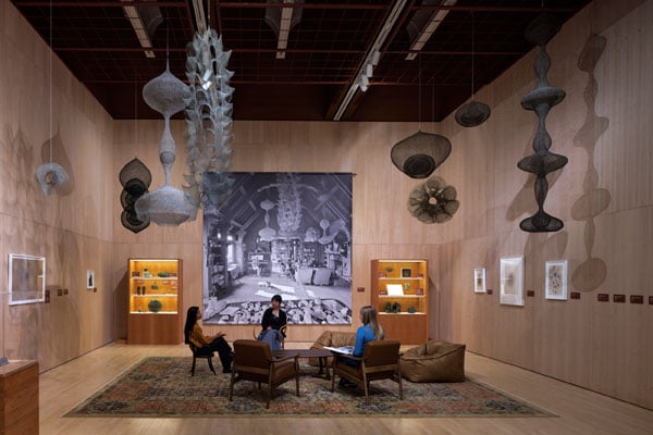 A gallery installed to resemble a living room features wire delicate sculptures dangling from the ceiling