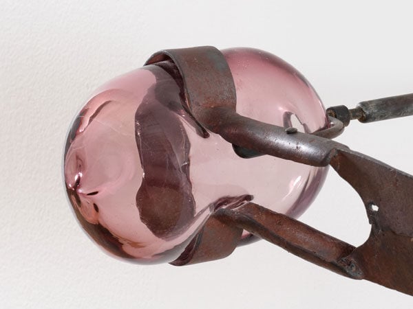 A detail image of a sculpture features bulbous pink glass held by iron tongs