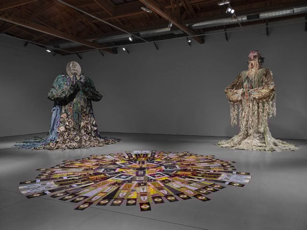 Two larger than life beings crafted from scraps of fabric rise over a gallery floor that contains a circular quilt.