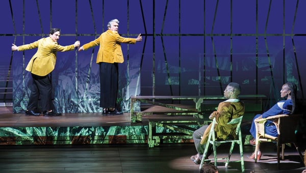 A scene on a stage shows two actors in bright yellow jackets