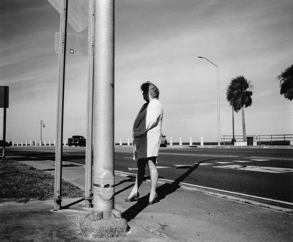 A black and white image shows a woman in heels and a long sweater standing in the shade of a telephone pole.