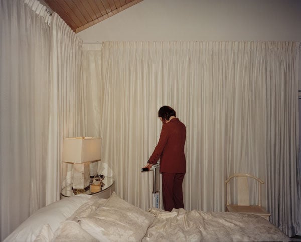 A woman in a burgundy suits has her back to the camera in a room surrounded by shiny white drapery and an equally shiny bedspread.