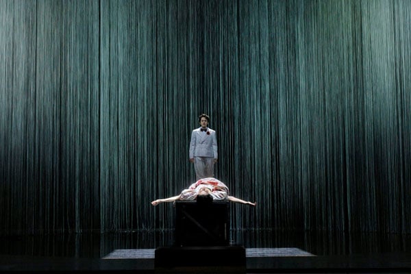 An opera singer in a man's suit stands over a woman laying on a platform, her dress bloodied