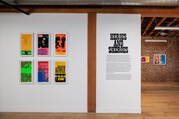 A view of an art gallery features bright printed posters and wall text that reads "heroes and sheroes"
